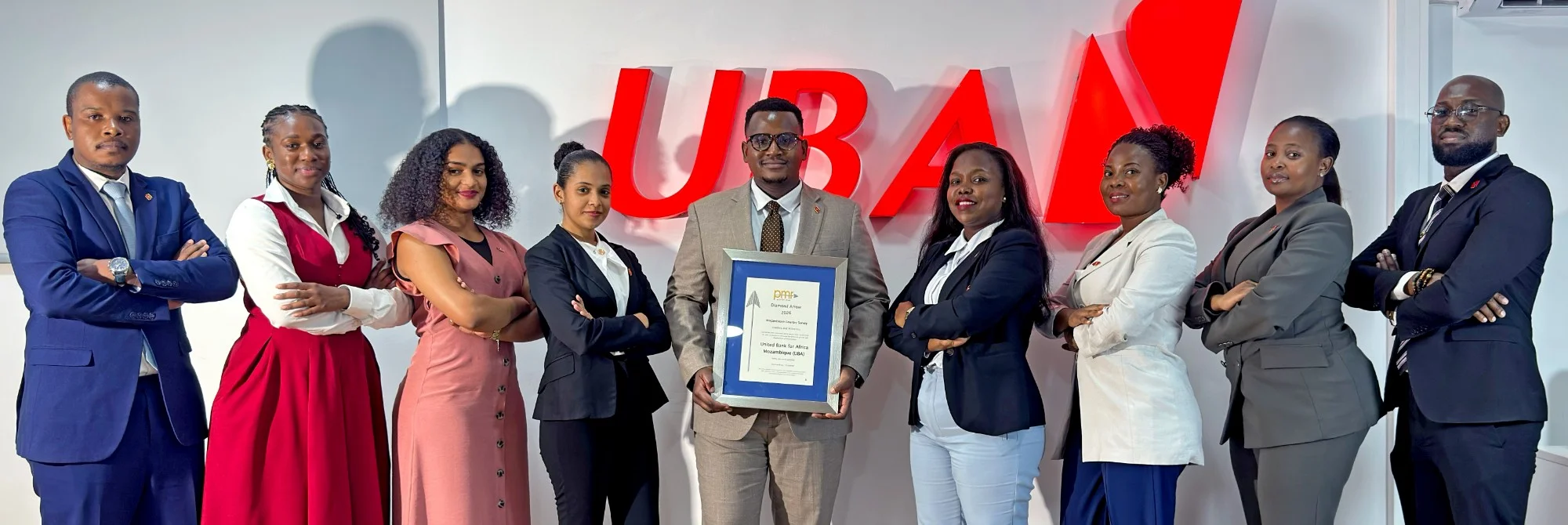 Grupo de oito pessoas de pé, de braços cruzados ou a segurar um quadro com um certificado, diante de um logótipo vermelho UBA numa parede branca