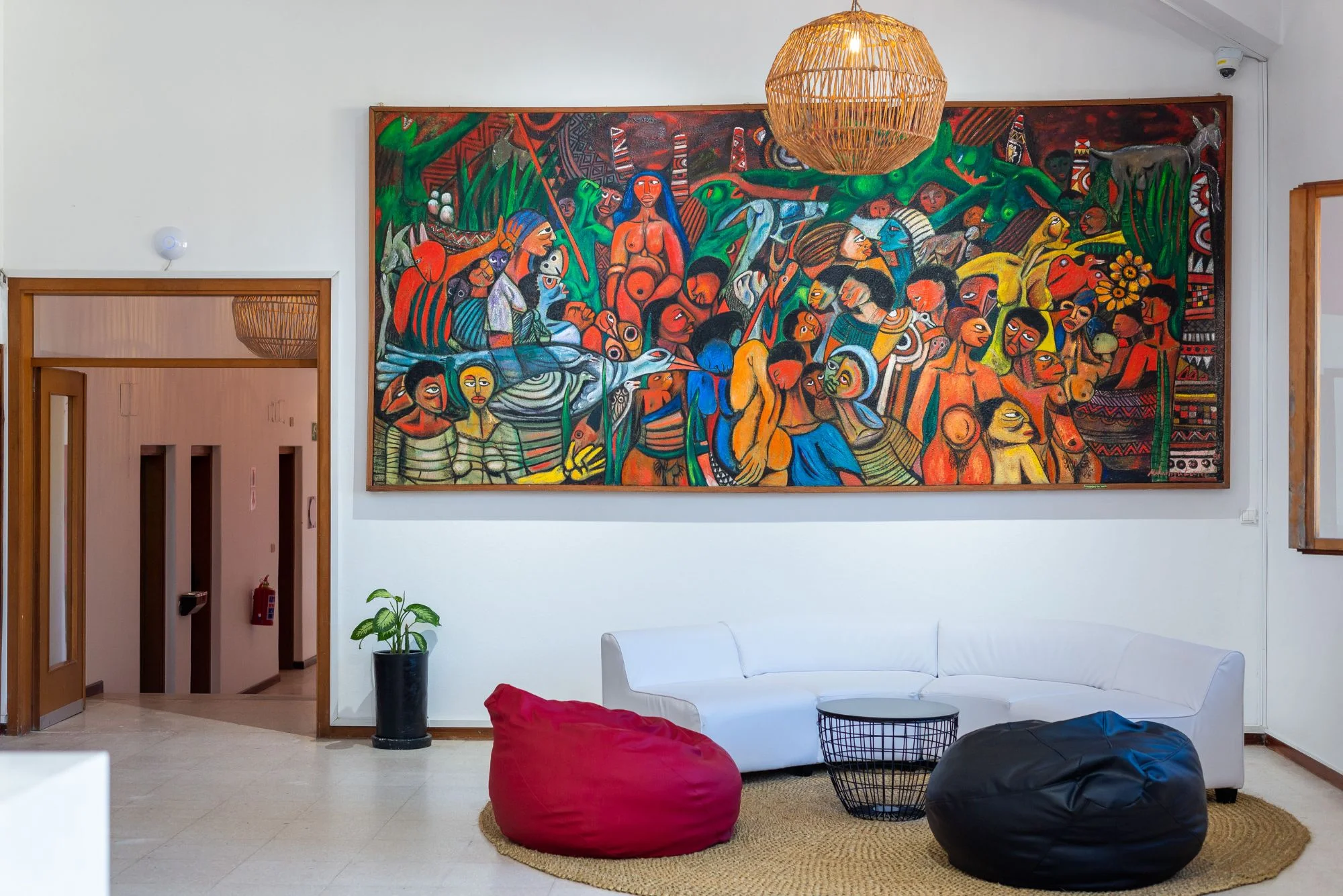 Sala de estar com uma grande pintura colorida de figuras na parede, sofá branco e pufes vermelho e preto sobre um tapete