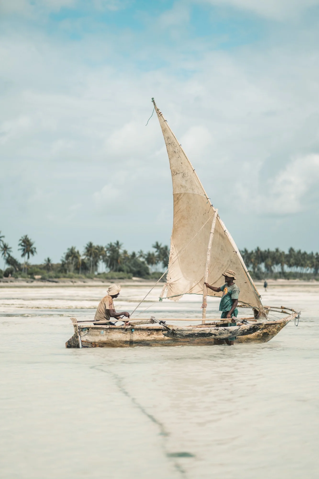 Dhow de madeira com vela triangular, em águas rasas e claras, com dois homens e palmeiras ao fundo