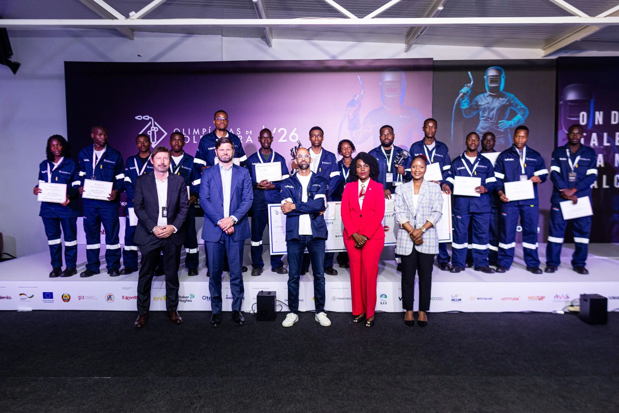 Grupo de participantes de uniforme azul-marinho com faixas refletoras, alguns com certificados na mão, e responsáveis de pé em primeiro plano, em frente a um palco com o logótipo das Olimpíadas de Soldadura '26