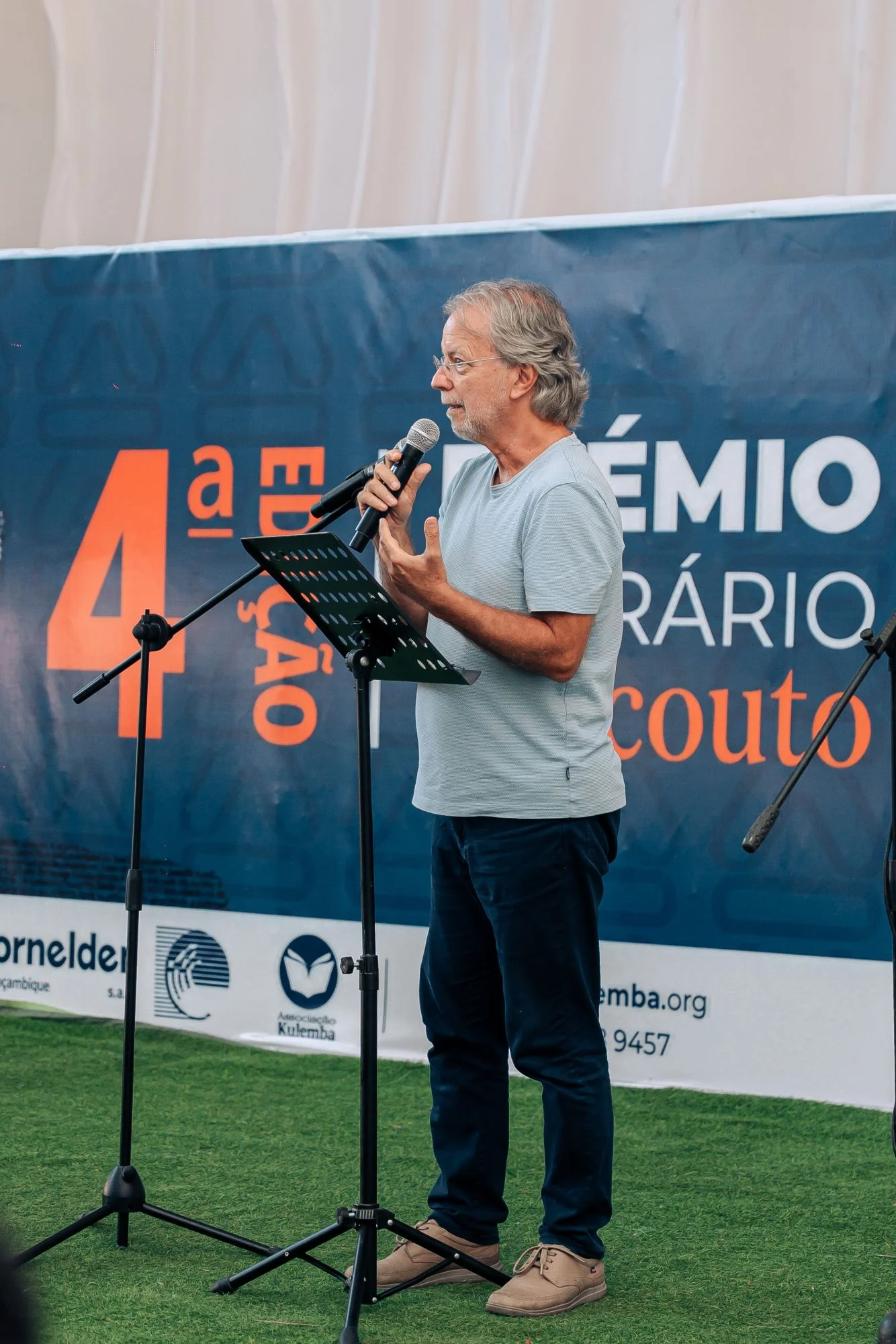 Homem de t-shirt cinzenta a falar ao microfone num púlpito, em frente a um painel azul com o logótipo do prémio