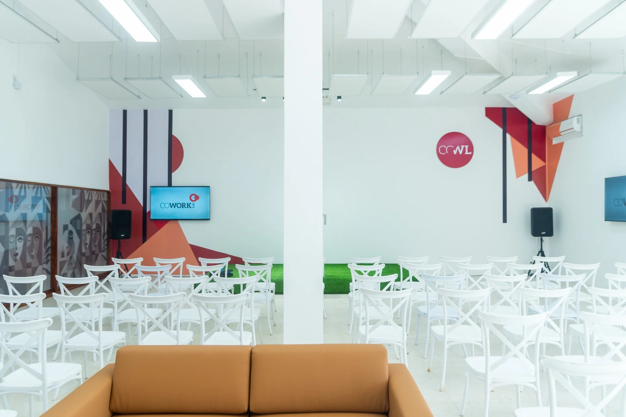 Sala de conferências com filas de cadeiras brancas viradas para um ecrã, uma pequena zona de relva verde e painéis geométricos vermelhos e laranja na parede