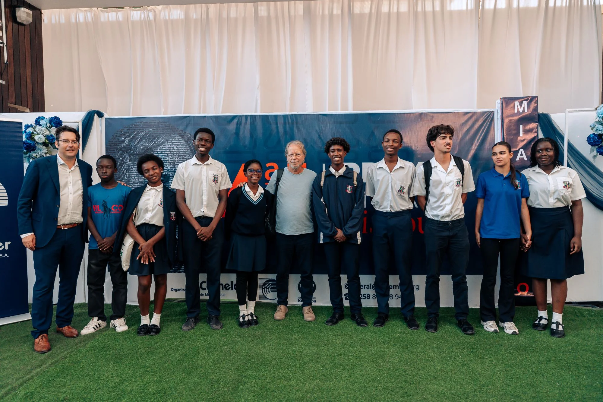 Grupo de pessoas de pé num relvado, a sorrir, incluindo jovens de uniforme escolar, em frente a um painel azul