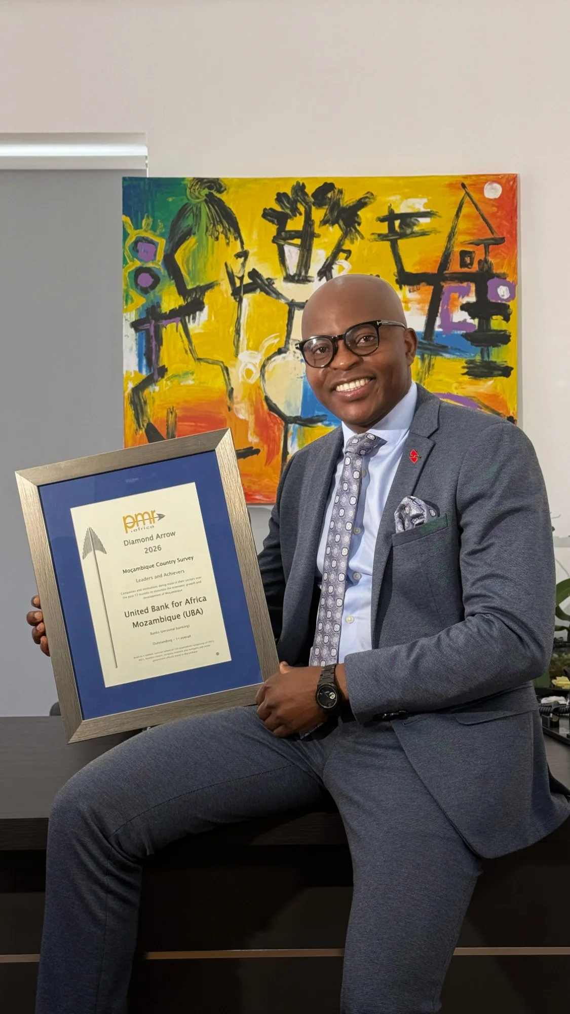 Homem sorridente, de fato cinzento e óculos, sentado num móvel de escritório a segurar um quadro com um certificado, diante de uma pintura colorida