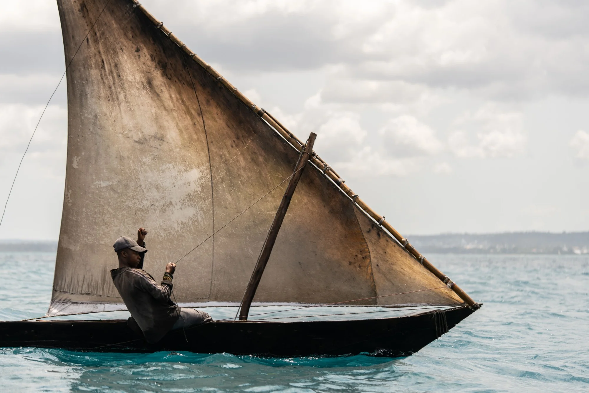 Barco de madeira com vela triangular de tons claros e um homem sentado a manobrar, num mar azul-acinzentado sob céu nublado