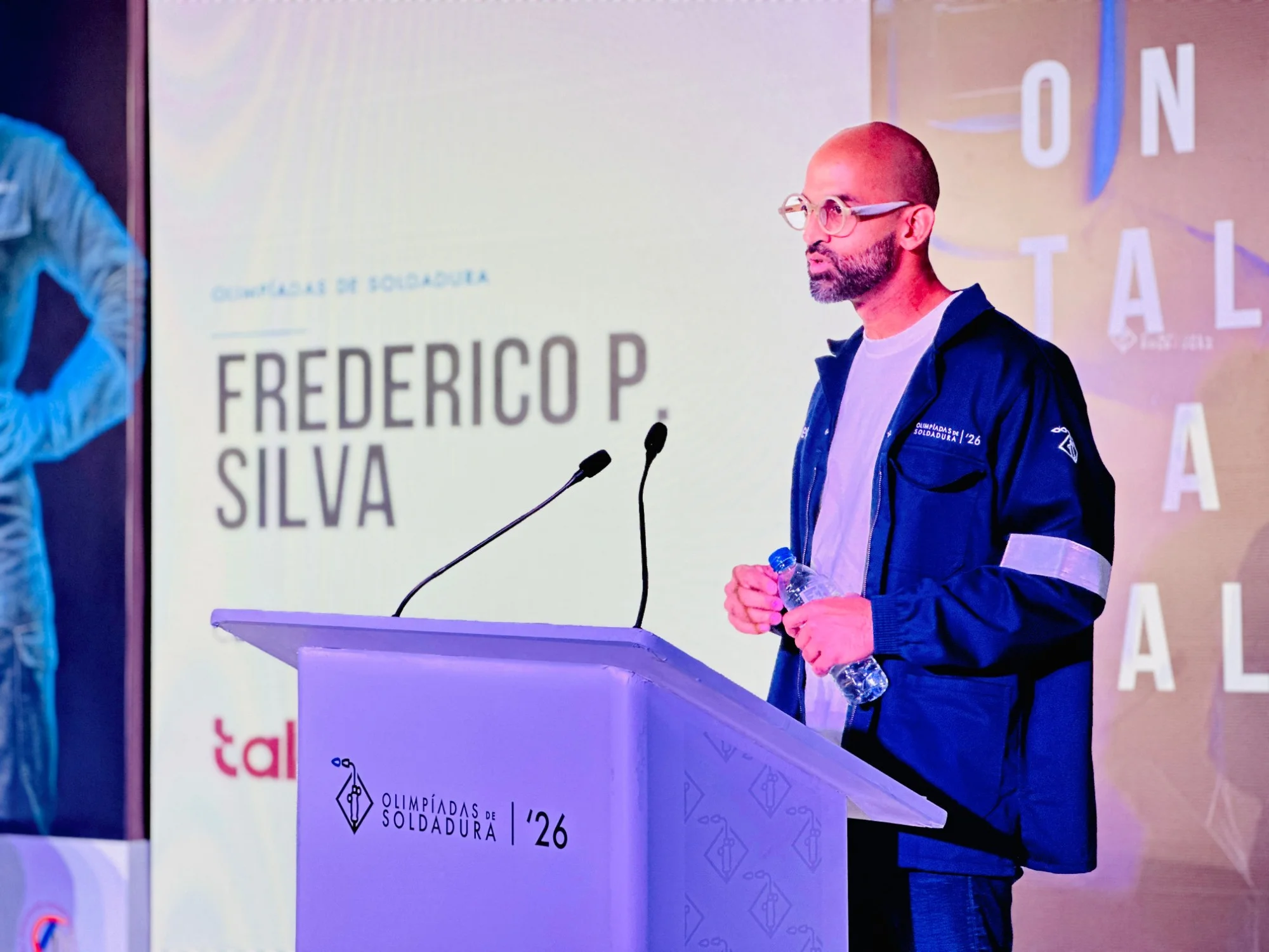 Homem de óculos e uniforme azul-marinho a falar ao microfone num púlpito, em frente a um ecrã com texto de identificação do orador