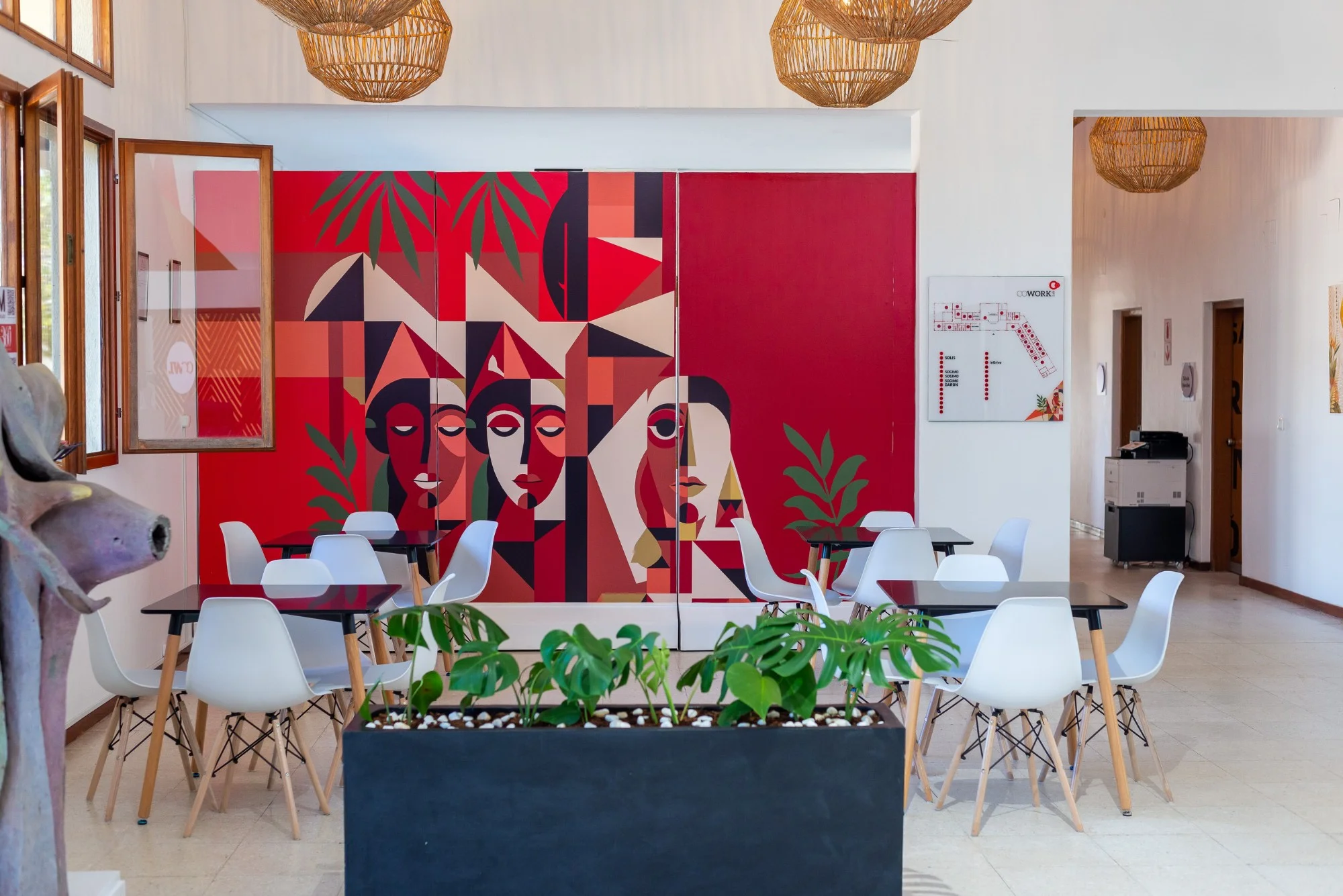 Espaço interior com parede vermelha pintada com figuras geométricas de rostos, mesas com cadeiras brancas e uma floreira com plantas em primeiro plano