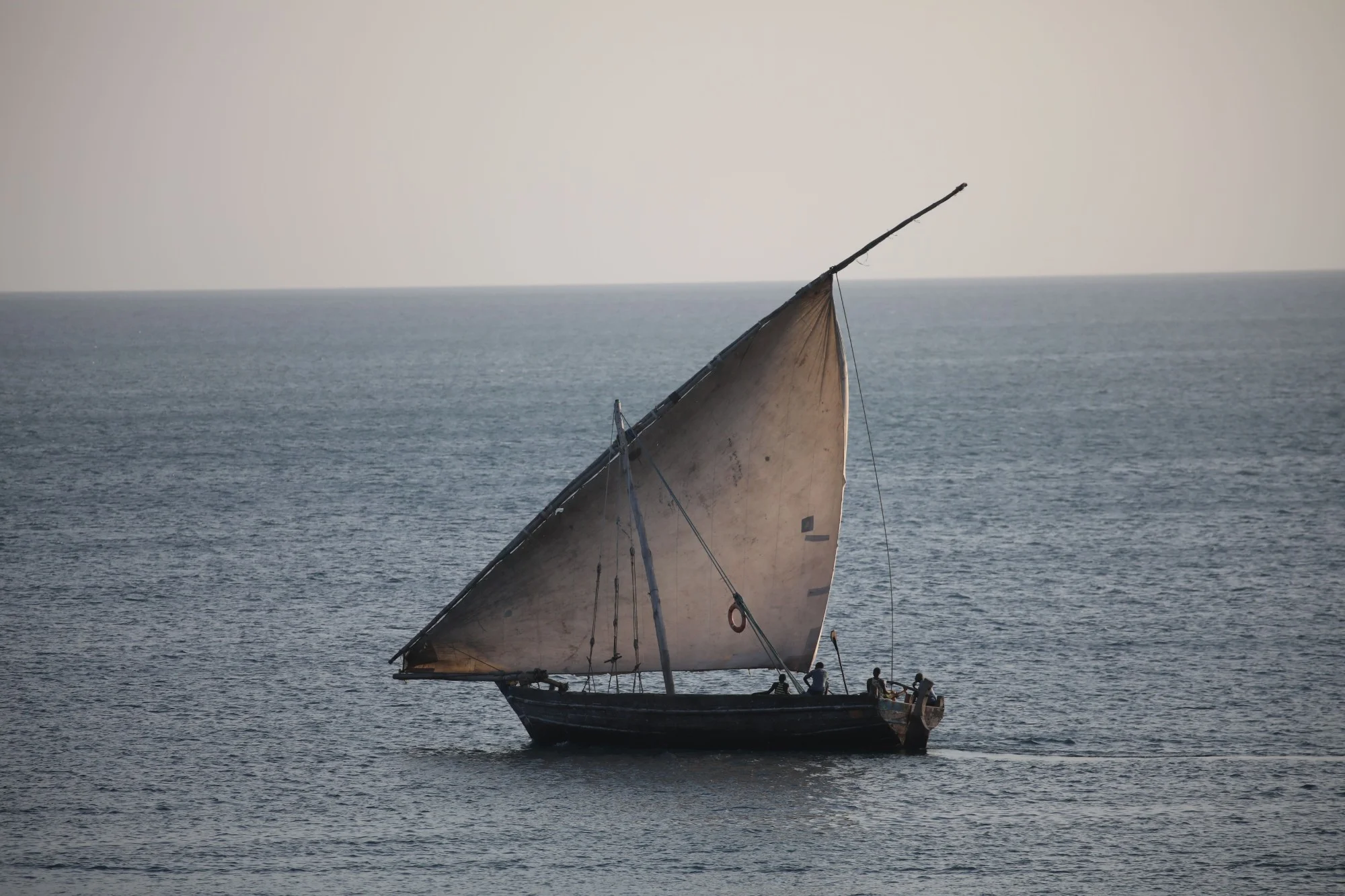 Dhow de madeira escura com vela triangular, a navegar em mar calmo, com tripulantes a bordo