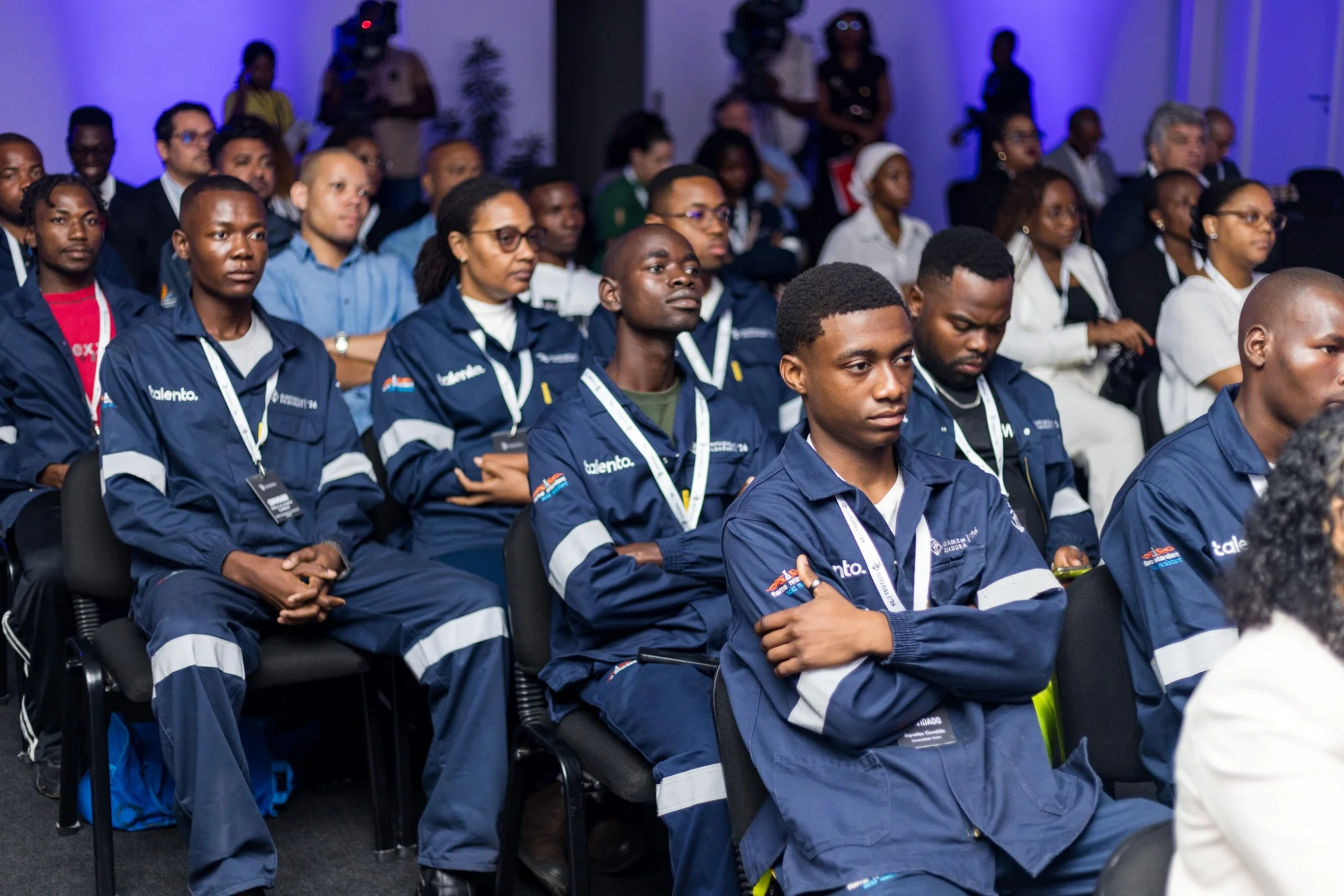 Plateia de participantes de uniforme azul-marinho sentados, atentos, numa sala com público atrás