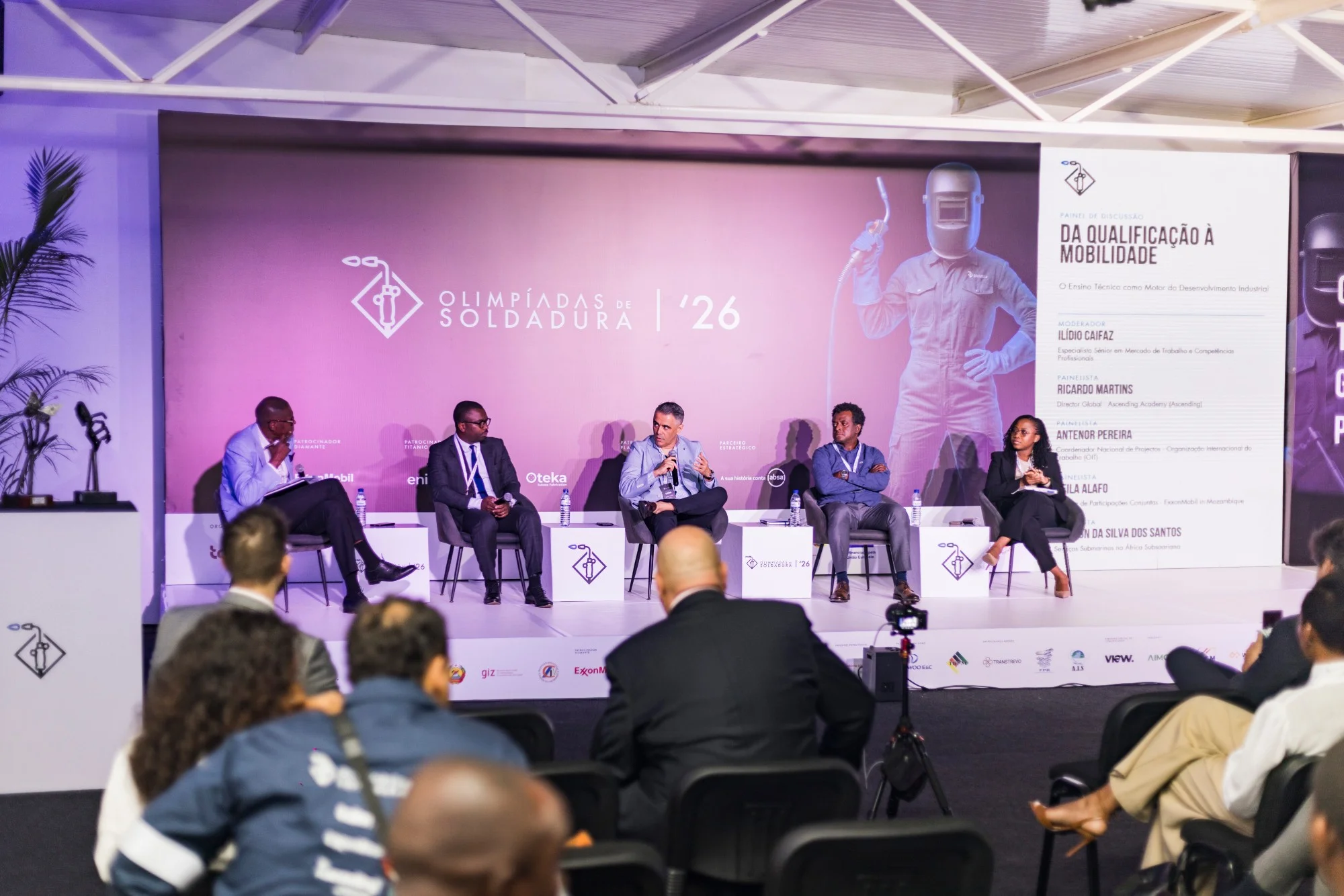 Painel de discussão com cinco participantes sentados num palco de fundo cor-de-rosa com logótipos, e o público de costas em primeiro plano